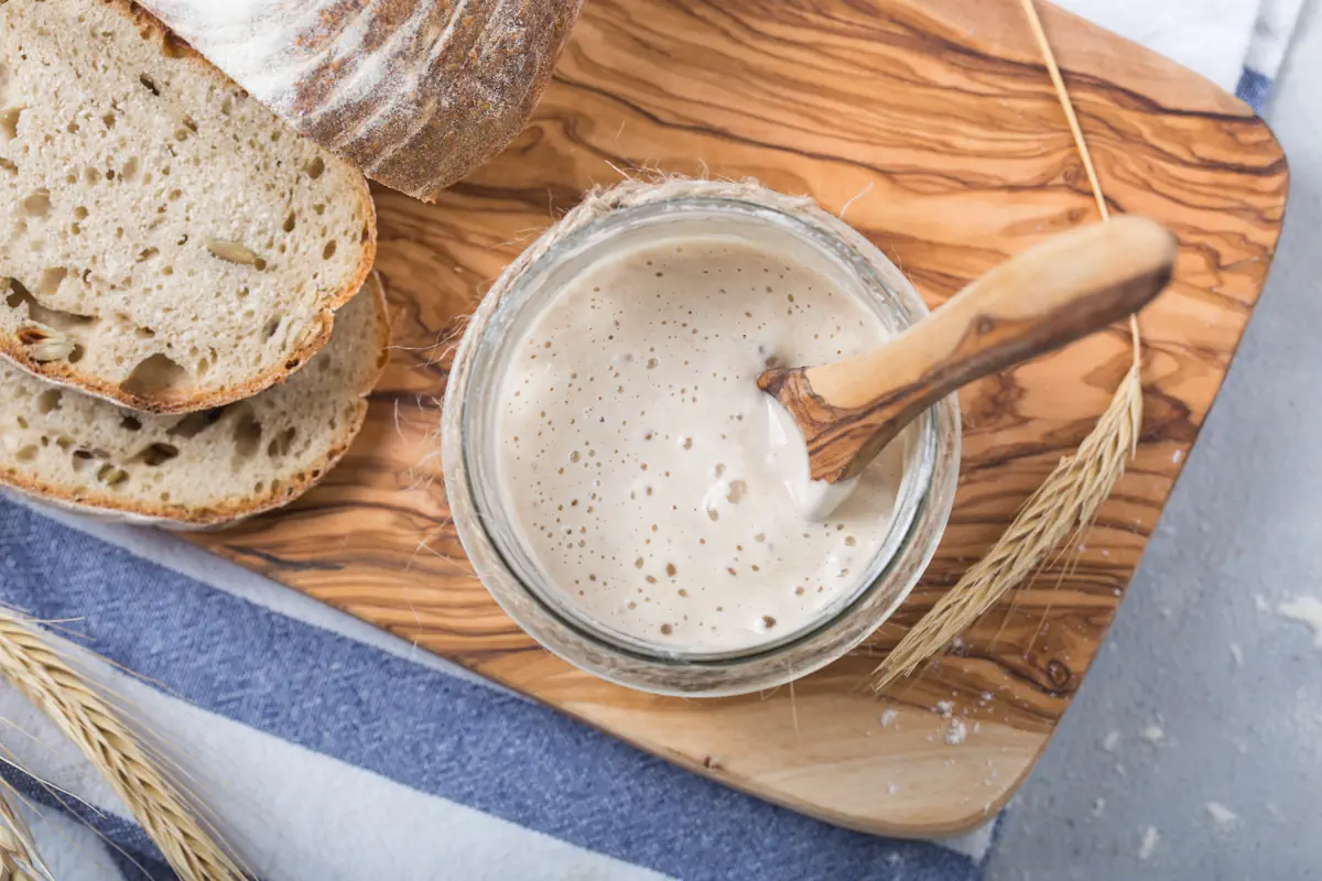 Un levain dans un bocal en verre posé sur une surface en bois près de morceaux de pain
